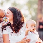 A tender family moment captured with a couple kissing and a baby holding bubbles outdoors.